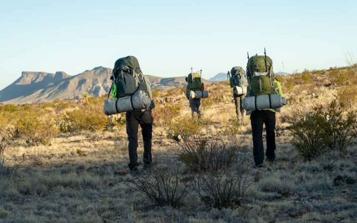 a group of students carrying backpacks hike through a desert landscape toward a mountain in the background